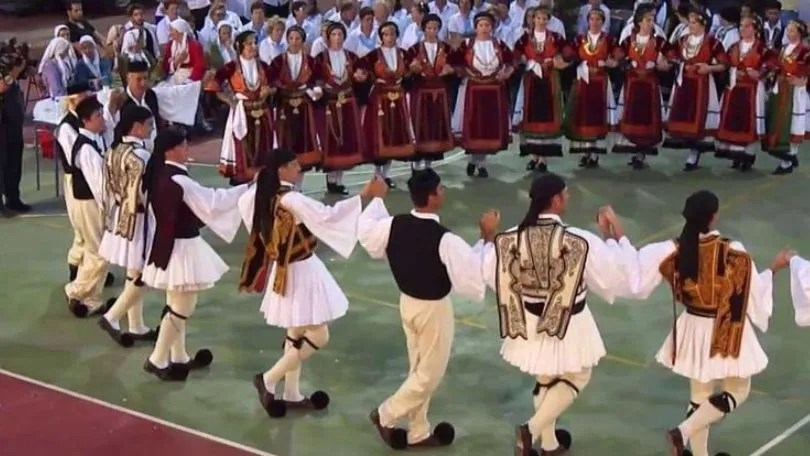 Group of around 20 mean and women dancing in traditional Greek costume to Greek music