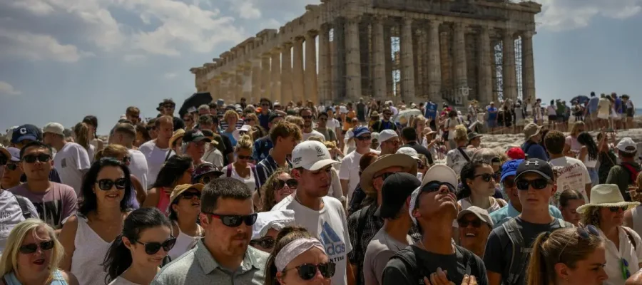 multitude of people visiting the Acropolis gathered at the entrance of the Acropolis in Athens, with many wearing hats and looking upwards towards the Parthenon under a partly cloudy sky.