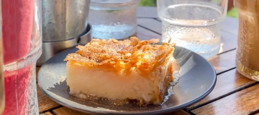 A slice of Galaktoboureko on a plate between galasses all sat on a wooden table. A slice of Galaktoboureko on a plate between galasses all sat on a wooden table. Served as part of the Street Food Tasting Tour in Athens